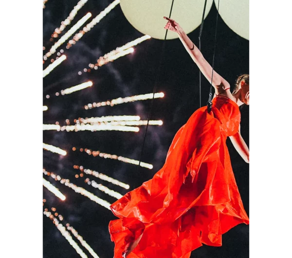 Aerial dancer in red dress flies through night sky with fireworks in the background