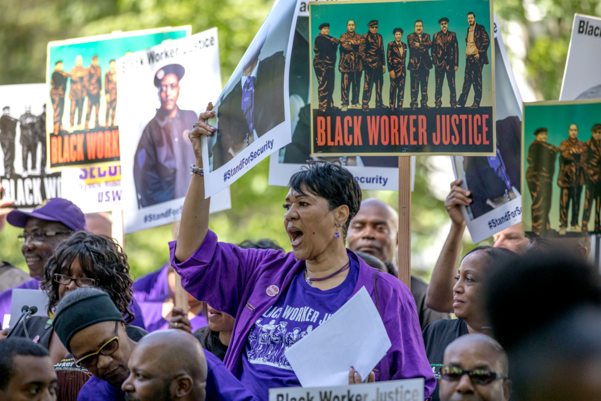 Photo of a Black woman in a purple shirt holding a poster in the middle of a crowd. Many other people posters are in the background