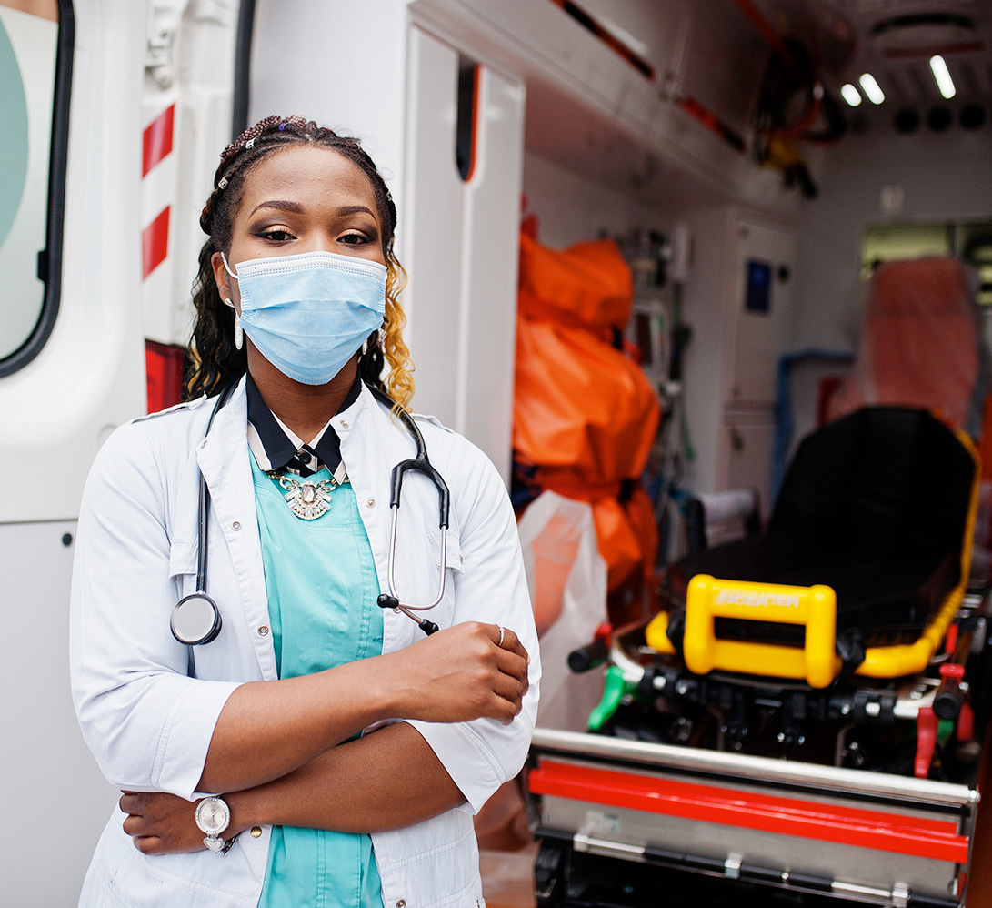 Black woman in a mask and doctor's lab coat standing in front of an open ambulance