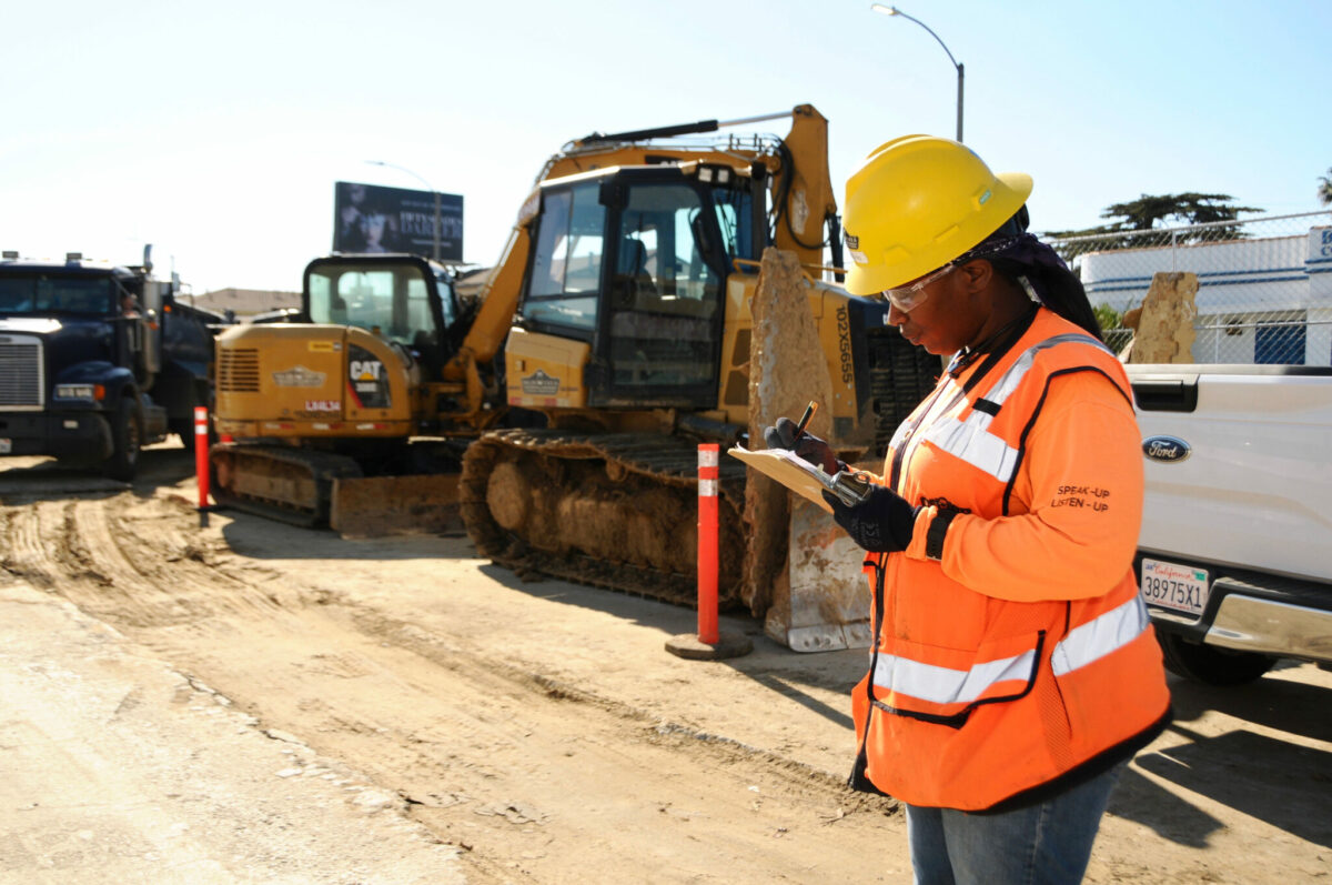Photo of a black woman construction worker standing in front of an excavator vehicle